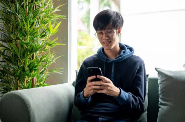 Kapüşonlu mutlu bir Asyalı genç, akıllı telefonuyla arkadaşlarıyla sohbet ederken evdeki kanepede dinleniyor. mesajları cevaplamak, internetten alışveriş yapmak, sosyal medyada kaydırmak, uygulamayı kullanmak