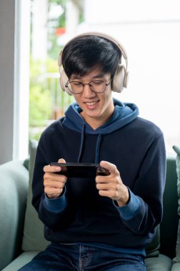 Kapüşonlu ve kulaklıklı genç, neşeli Asyalı bir adam evdeki kanepede mobil oyun oynamaktan zevk alıyor. İnsanlar, eğlence, kablosuz teknoloji