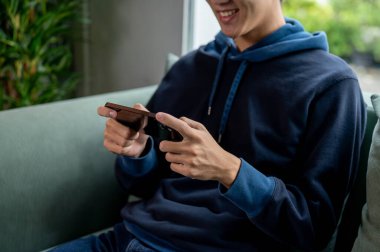 Mavi kapüşonlu genç, neşeli Asyalı bir adamın kırpılmış görüntüsü evdeki kanepede mobil bir oyun oynuyor. İnsanlar, eğlence, kablosuz teknoloji