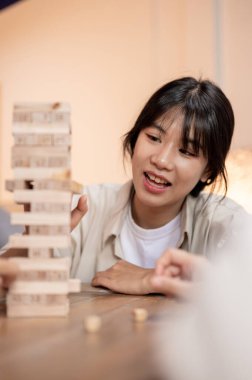 Genç, neşeli bir Asyalı kız arkadaşlarıyla evde Jenga oynamaktan zevk alıyor, tahta bir bloğu kaldırmaya ve ahşap bir kule inşa etmeye odaklanıyor. Oyun masası, boş zaman, eğlence aktivitesi