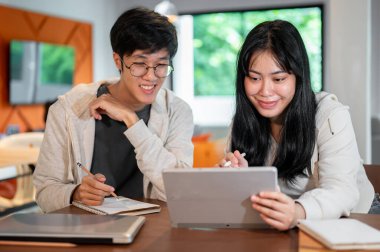İki pozitif Asyalı genç üniversite öğrencisi, bir kafede veya kütüphanede birlikte çalışma alanında dijital tablet üzerine online bir ders çalışıyorlar. Eğitim ve kablosuz teknoloji kavramları