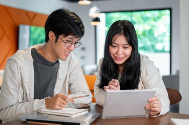 İki pozitif Asyalı genç üniversite öğrencisi, bir kafede veya kütüphanede birlikte çalışma alanında dijital tablet üzerine online bir ders çalışıyorlar. Eğitim ve kablosuz teknoloji kavramları