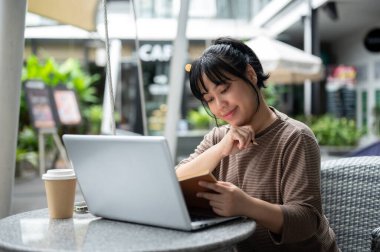Pozitif Asyalı bir kadın bir kafede uzaktan çalışıyor, açık bir masada oturuyor, dizüstü bilgisayarını kullanıyor ve kitap okuyor veya bir kitaba not alıyor. insanlar ve şehir hayatı kavramları