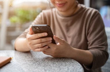 Açık hava masasında dinlenirken telefonuna kayan Asyalı bir kadının kırpılmış görüntüsü. Sohbet, online alışveriş, mesajlaşma, cep telefonu uygulaması kullanma, e-postaları kontrol etme