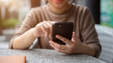 Açık hava masasında dinlenirken telefonuna kayan Asyalı bir kadının kırpılmış görüntüsü. Sohbet, online alışveriş, mesajlaşma, cep telefonu uygulaması kullanma, e-postaları kontrol etme