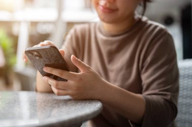 Açık hava masasında dinlenirken telefonuna kayan Asyalı bir kadının kırpılmış görüntüsü. Sohbet, online alışveriş, mesajlaşma, cep telefonu uygulaması kullanma, e-postaları kontrol etme