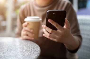 Akıllı telefonunu kullanan, kahve içen, şehirdeki bir kahve dükkanının açık hava masasında oturan bir kadının kırpılmış görüntüsü. İnsanlar ve kablosuz teknoloji kavramları