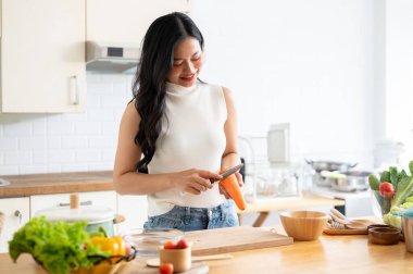 Çekici, çekici bir Asyalı kadın mutfakta yemek pişiriyor, mutfak adasında bıçakla havuç soyuyor, ona sağlıklı bir kahvaltı hazırlıyor. Ev yemeği, ev hayatı, sağlıklı yaşam tarzı.