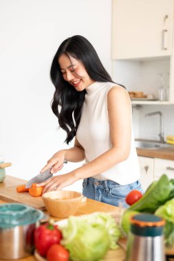 Çekici, mutlu bir Asyalı kadın kesim tahtasının üzerinde havuç doğruyor, mutfakta yemek pişiriyor, sağlıklı bir kahvaltı hazırlıyor. Ev yemeği, sağlıklı yaşam tarzı, ev hayatı.