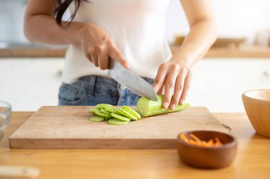 Mutfak masasında salatalığı bıçakla doğrayan, yemek pişiren bir kadının kırpılmış görüntüsü. Sağlıklı yemek, ev yemeği, sağlıklı beslenme, ev hayatı.