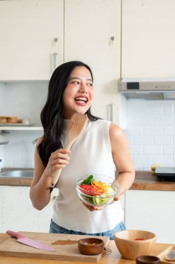 Çekici bir Asyalı kadın mutfakta elinde bir kase salata ve ahşap bir spatulayla mutfakta yemek pişirmekten zevk alıyor. Ev yemekleri ve sağlıklı beslenme konseptleri.