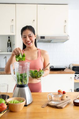 Sağlıklı, formda, spor giyimli genç bir Asyalı kadın sağlıklı yeşil smoothie 'sini mutfakta blender ile yapıyor. Sağlıklı yiyecekler, sağlıklı yaşam tarzı, kilo verme tarifi, ev yemekleri.