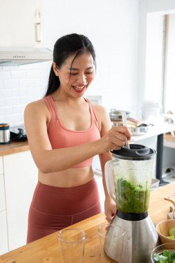 Güçlü, sağlıklı bir Asyalı kadın spor giyim sektöründe mutfakta blender ile sağlıklı yeşil bir smoothie hazırlıyor. Antrenmandan sonra ona sağlıklı bir içecek hazırlıyor. sağlıklı yaşam tarzı ve ev yemeği kavramları