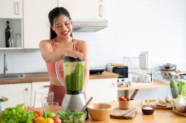 Spor giyimli, çekici, zayıf Asyalı bir kadın yeşil meyve suyunu mutfakta blender ile yapıyor. Egzersiz yaptıktan sonra sağlıklı enerji içeceği yapıyor. sağlıklı yaşam tarzı, sağlıklı yaşam, ev hayatı