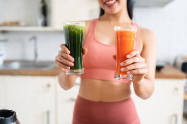 Çekici ve sağlıklı bir Asyalı kadının yakın plan görüntüsü. Pembe spor kıyafetleri içinde. Mutfakta yeşil ve turuncu meyve suyu bardakları tutuyor. Sağlıklı yemek, diyet, refah, beslenme