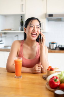 Neşeli, çekici bir Asyalı kadın telefonda konuşmaktan ve mutfaktaki bir mutfak adasında smoothie içmekten zevk alıyor. sağlıklı yaşam tarzı kavramı