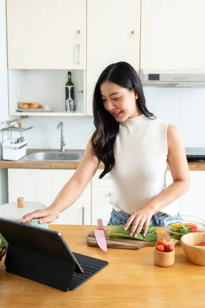 stock image A gorgeous young Asian woman using her digital tablet while cooking in the kitchen, following online recipe on the internet. home cooking and lifestyle concepts