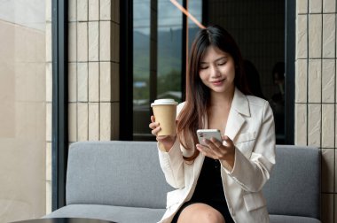 Güzel, mutlu bir Asyalı iş kadını bir kafede dinleniyor, akıllı telefonuyla kahve içiyor. metin okumak, mesajları kontrol etmek, sohbet cevaplamak, sosyal medyada videolar izlemek