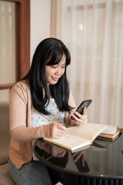 Güzel, pozitif Asyalı bir kadın akıllı telefonunu kullanıyor, defterine not alıyor, kapalı bir masada oturuyor..