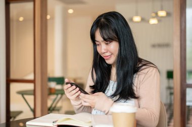Olumlu, çekici Asyalı bir kadın kitaplarıyla bir kafede otururken akıllı telefonunu kullanıyor. telefonda kaydediliyor, sosyal medya, sohbet