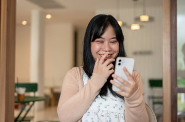 Neşeli bir Asyalı kadın akıllı telefonunda mesajlar okurken ve bir kafede dinlenirken gülüyor. sohbet, sosyal medya, insanlar ve yaşam tarzları kavramları