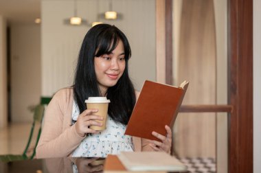 Güzel, pozitif Asyalı bir kadın güzel bir kahve dükkanında kitap okurken sıcak bir şeyler içiyor. İnsanlar, eğlence ve yaşam tarzı kavramları