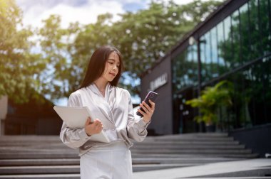 Yakışıklı, başarılı Asyalı bir iş kadını şık bir ofis elbisesi içinde elinde dosya klasörüyle merdivenlerden inerken akıllı telefonundan gelen mesajları kontrol ediyor..
