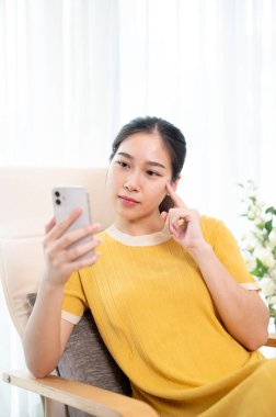 Düşünceli bir Asyalı kadın akıllı telefonuyla evde koltukta dinlenirken mesajlar okuyor. İnsanlar ve kablosuz teknoloji kavramları
