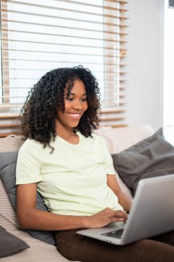 Mutlu bir genç siyahi kadın evdeki bir kanepede dizüstü bilgisayarıyla çalışıyor. Uzaktan çalışmak, evden çalışmak, internette sörf yapmak, sohbet etmek.