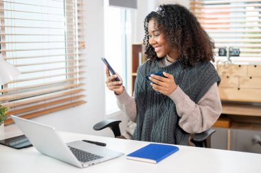 Samimi örgü süveter giyen, kulaklık takan çekici siyah bir kadın, ofisinde sabah kahvesini içerken telefonuna kayıyor..