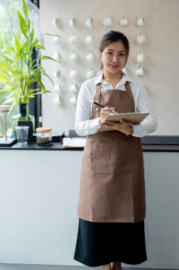 Kahverengi önlüklü çekici, gülümseyen Asyalı genç bir bayan garson ya da barista elinde menüyle minimal beyaz bir kahve dükkanında duruyor. Küçük işletme, işletme sahibi, çalışan