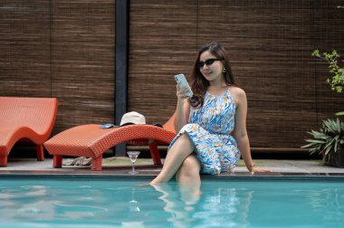 Güzel mavi elbiseli ve güneş gözlüklü zarif, kendine güvenen Asyalı bir kadın havuz kenarında oturuyor ve akıllı telefonunu kullanıyor. Yaz tatili, lüks yaşam tarzı, eğlence.