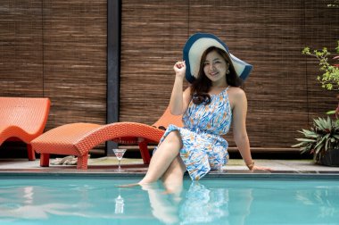 Güzel bir yaz elbisesi ve plaj şapkası içinde zarif, mutlu bir Asyalı kadın havuz kenarında oturuyor ve yaz tatilinin tadını çıkarıyor. Lüks yaşam tarzı, keyif, rahatlama.