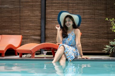 Güzel bir yaz elbisesi ve plaj şapkası içinde zarif, mutlu bir Asyalı kadın havuz kenarında oturuyor ve yaz tatilinin tadını çıkarıyor. Lüks yaşam tarzı, keyif, rahatlama.