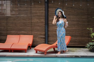 Güzel bir yaz elbisesi ve plaj şapkası giymiş zarif, kendine güvenen Asyalı bir kadın elinde votka martini bardağıyla havuz kenarında yürüyor. Otel havuz salonu, yaz tatili, lüks yaşam tarzı.