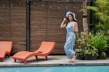 Güzel bir yaz elbisesi ve plaj şapkası giymiş zarif, kendine güvenen Asyalı bir kadın elinde votka martini bardağıyla havuz kenarında yürüyor. Otel havuz salonu, yaz tatili, lüks yaşam tarzı.