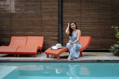Güzel bir yaz elbisesi içinde güzel bir Asyalı kadın havuz kenarındaki şezlongda oturuyor, otelde tatilin tadını çıkarıyor..