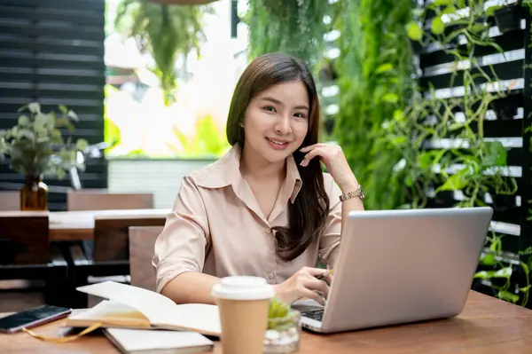 Çekici bir Asyalı iş kadını bir kafede uzaktan çalışıyor, açık bir masada dizüstü bilgisayarıyla oturuyor, kameraya gülümsüyor..