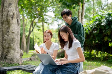 Bir grup mutlu Asyalı genç üniversite öğrencisi projelerini tartışıyorlar, fikirlerini paylaşıyorlar, dizüstü bilgisayar kullanıyorlar, parktaki bankta birlikte çalışıyorlar. Eğitim ve arkadaşlık kavramları
