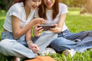 İki güzel Asyalı genç bayan arkadaşın kesilip biçilmiş görüntüsü parkta çimenlerde dinlenirken akıllı telefondan konuşmaktan ve bir şeyler izlemekten keyif alıyorlar. dostluk ve yaşam tarzları