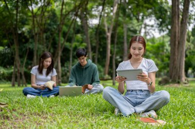 Genç mutlu bir Asyalı kız üniversite öğrencisi kampüs parkında otururken dijital tabletini okuyor. Eğitim ve kablosuz teknoloji kavramları