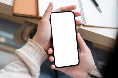Kapalı bir masada kitaplarıyla oturan ve akıllı telefonunu kullanan bir kadının yakın çekim görüntüsü. Beyaz ekran modeli, insan ve kablosuz teknolojisi olan bir akıllı telefon.