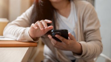 Rahat giysiler içinde Asyalı bir kadın minimalist bir kahve dükkanında oturmuş, akıllı telefonunu kullanıyor, biriyle sohbet ediyor ya da telefonunu kaydırıyor..