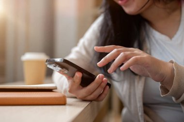 Rahat giysiler içinde Asyalı bir kadın minimalist bir kahve dükkanında oturmuş, akıllı telefonunu kullanıyor, biriyle sohbet ediyor ya da telefonunu kaydırıyor..
