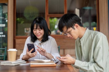 İki mutlu Asyalı genç arkadaş, bir erkek ve bir kadın, bir kafede oturmuş, telefonlarını kullanıyorlar. sohbet, uygulama kullanımı, telefonda kaydırma, çevrimiçi blog okuma