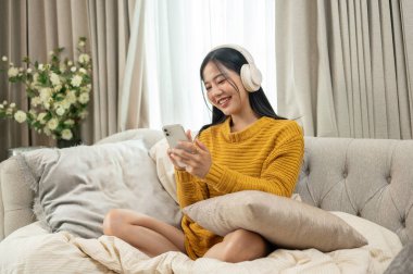 Hoş sarı kazaklı, güzel, pozitif Asyalı bir kadın akıllı telefonuyla çalma listesinden bir şarkı seçiyor ve evdeki bir kanepede müzik dinlemenin keyfini çıkarıyor..