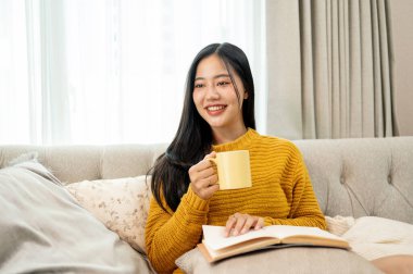 Çekici bir Asyalı kadın oturma odasındaki kanepede kitap okurken kahve içiyor, dışarıdaki manzaraya bakıyor ve hayal kuruyor. Boş zaman, yaşam tarzı, ev hayatı.