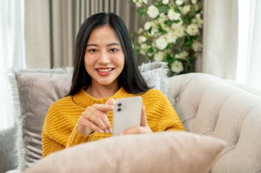 Hoş örgü süveter giymiş çekici bir Asyalı kadın evdeki kanepede dinleniyor, akıllı telefonunu tutuyor ve kameraya gülümsüyor. İnsanlar, teknoloji ve ev hayatı