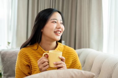 Güzel, çekici, örgü örülü bir kazak giyen Asyalı bir kadın elinde kahve fincanı, oturma odasında bir kanepede oturuyor, gülümsüyor ve hayal kuruyor..