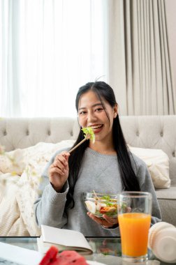Olumlu, çekici, genç bir Asyalı kadın, dizüstü bilgisayarı üzerinde çalışırken oturma odasındaki kahve masasında sağlıklı salata yemekten zevk alıyor. Evden çalışmak, boş zaman, ev hayatı.